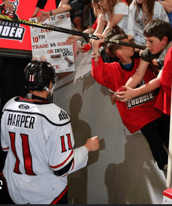 At a sports game, a hockey player wearing a jersey labeled Harper greets excited young fans behind the glass, who are reaching out with jerseys, sticks, and signs for autographs.